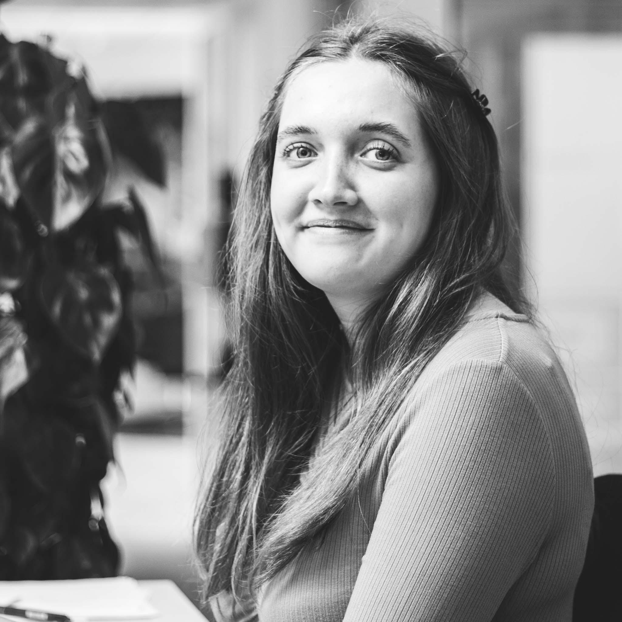 Sandra sidder ved skrivebord med plante i baggrund.